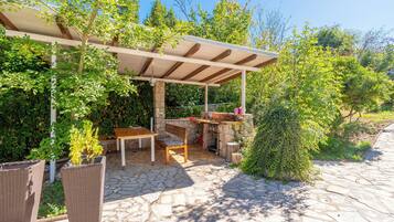 Eine Terrasse im Freien mit einer Pergola, Steinboden, einem Tisch, Stühlen und einer kleinen Küchenzeile.