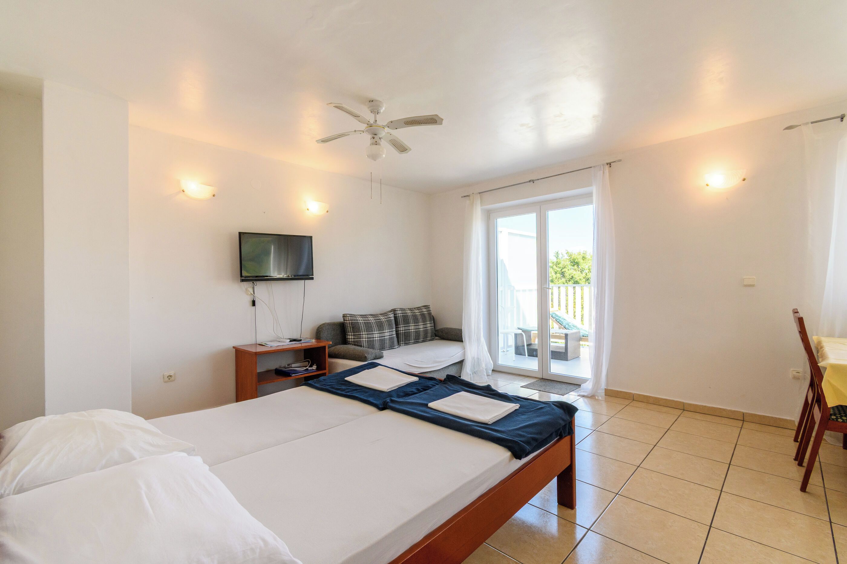 A hotel room with a bed, a TV, a ceiling fan, and a view of the outside.