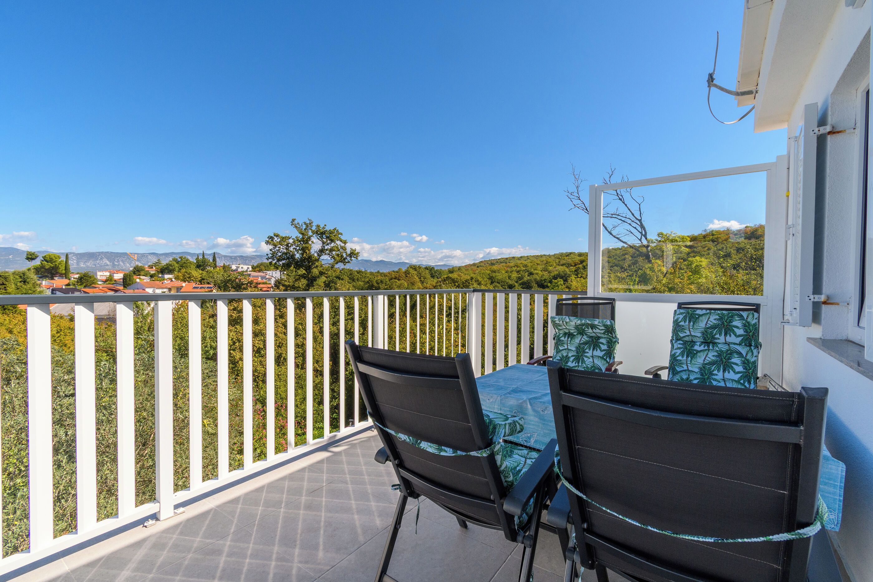 A balcony with two chairs and a table, overlooking a scenic landscape with trees and distant mountains.
