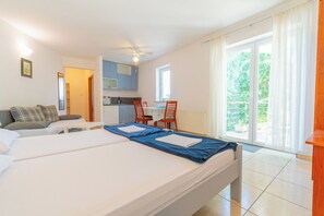 A hotel room with a bed, a sofa, a dining table with chairs, a kitchen area, and a balcony with a view of greenery. - Apartments Vlado (Omisalj)