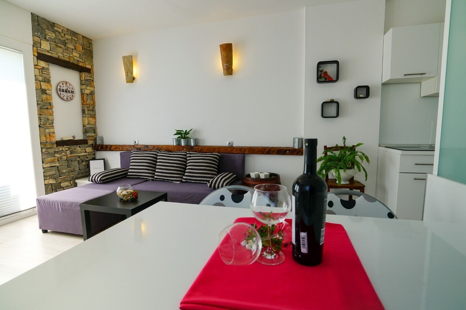 A modern living room with a stone fireplace, a sofa with striped cushions, a dining table with a red tablecloth, and a bottle of wine.