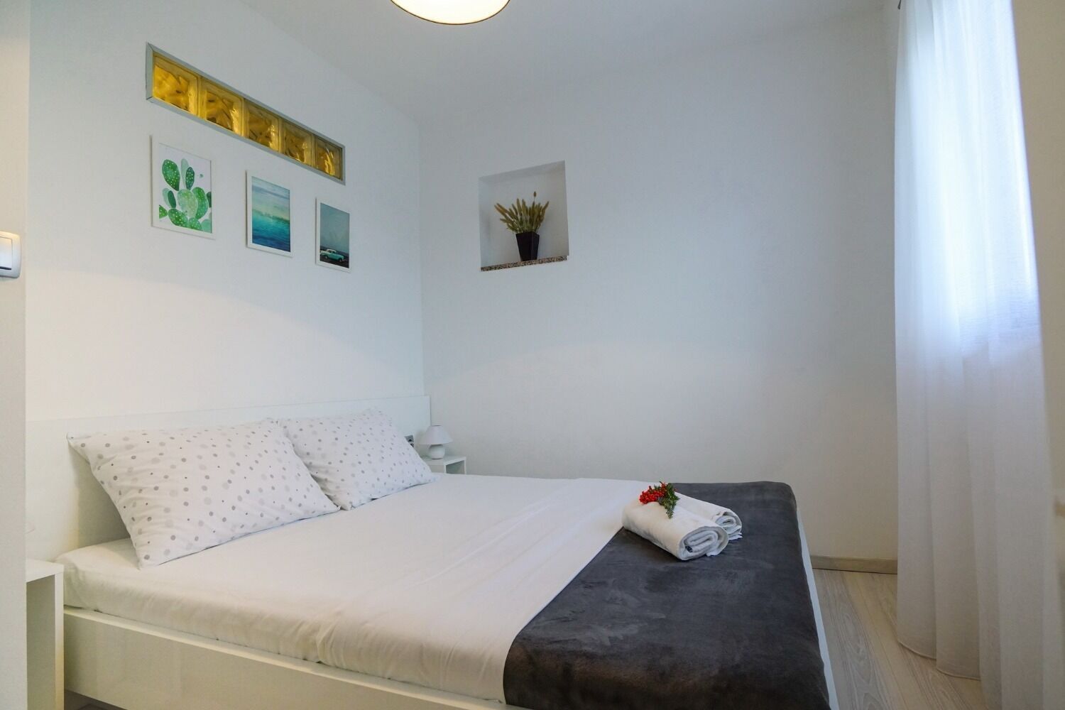 A neatly made bed with white bedding and a dark grey blanket, a small bedside table with a lamp, and a wall-mounted shelf with a potted plant.