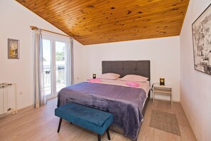 A bedroom with a wooden ceiling, a bed with a purple blanket, a bench, a nightstand with a lamp, and a window with curtains. - Apartments Nadi (Hvar)