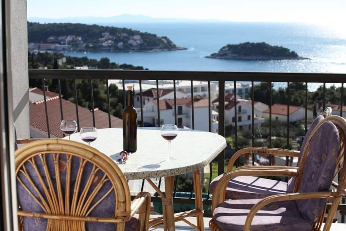 Ein Balkon mit Korbstühlen und einem Tisch, mit Blick auf die Küste, Häuser und ein Gewässer.