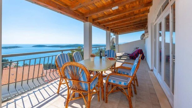En overdækket udendørs terrasse med et bord og stole, der ud til et smukt kig over havet og bakker.