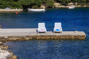 Due poltrone bianche con asciugamani su un molo in pietra che si protende su un corpo d'acqua, con barche e vegetazione sullo sfondo.