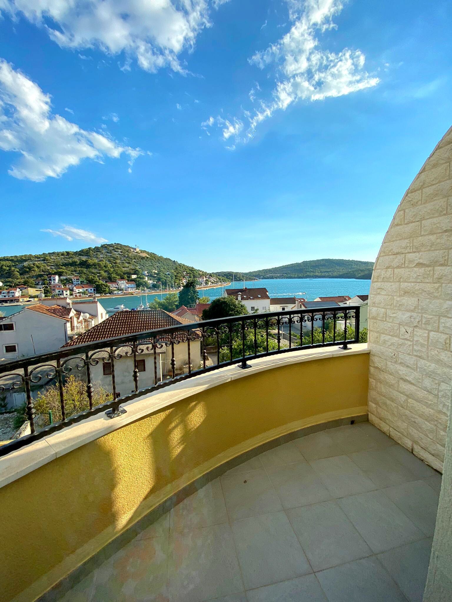 A balcony with a yellow wall and a view of a coastal town, including houses and a body of water.