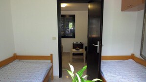 Ein Zimmer mit zwei Einzelbetten, einer Holztür, einem kleinen Tisch mit einer Pflanze und einem Fenster mit Jalousien.