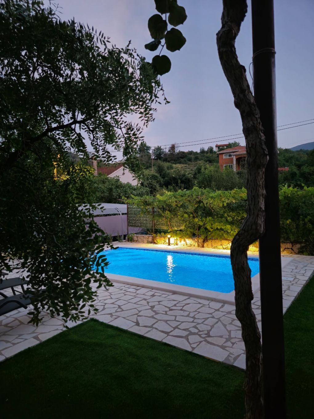 Una piscina con bordo piastrellato, circondata dal verde e con una casa sullo sfondo.