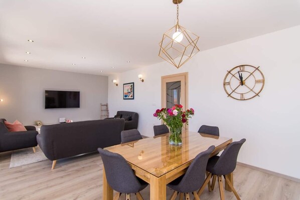 Un salon moderne avec une table à manger en bois, des canapés gris, un téléviseur fixé au mur et une horloge décorative.