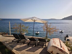 A seaside patio with lounge chairs and an umbrella, overlooking the sea with boats and a boat on the shore. - House Mirka (Rogoznica)