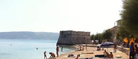 A beach with people sunbathing and swimming, a stone wall, and a building in the background.