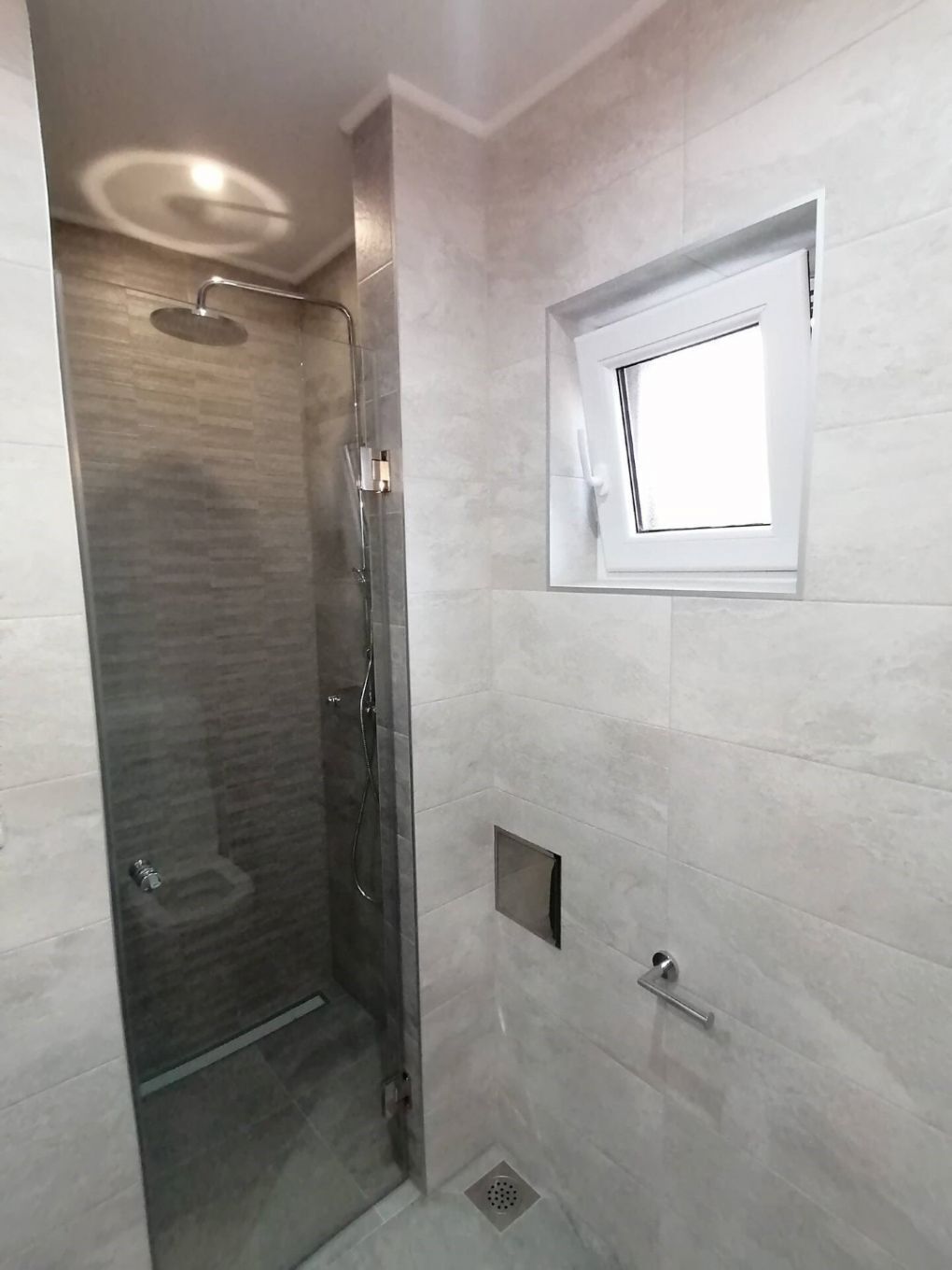 A modern bathroom with a glass-enclosed shower, a toilet, and a window above the sink.