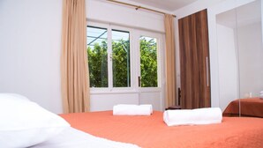 A hotel room with two beds, a large window with curtains, and a view of greenery outside. - Apartment Anka (Kastela)
