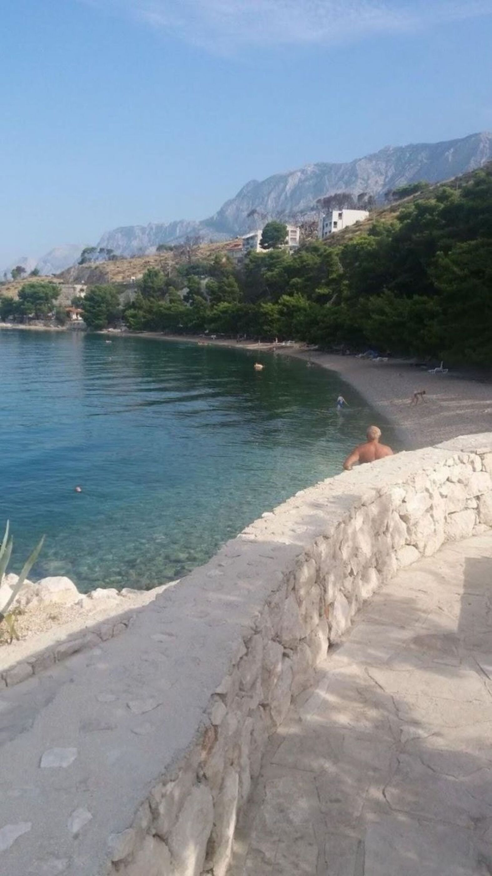 Paesaggio costiero con un muro di pietra, acqua blu cristallina e edifici su una collina.