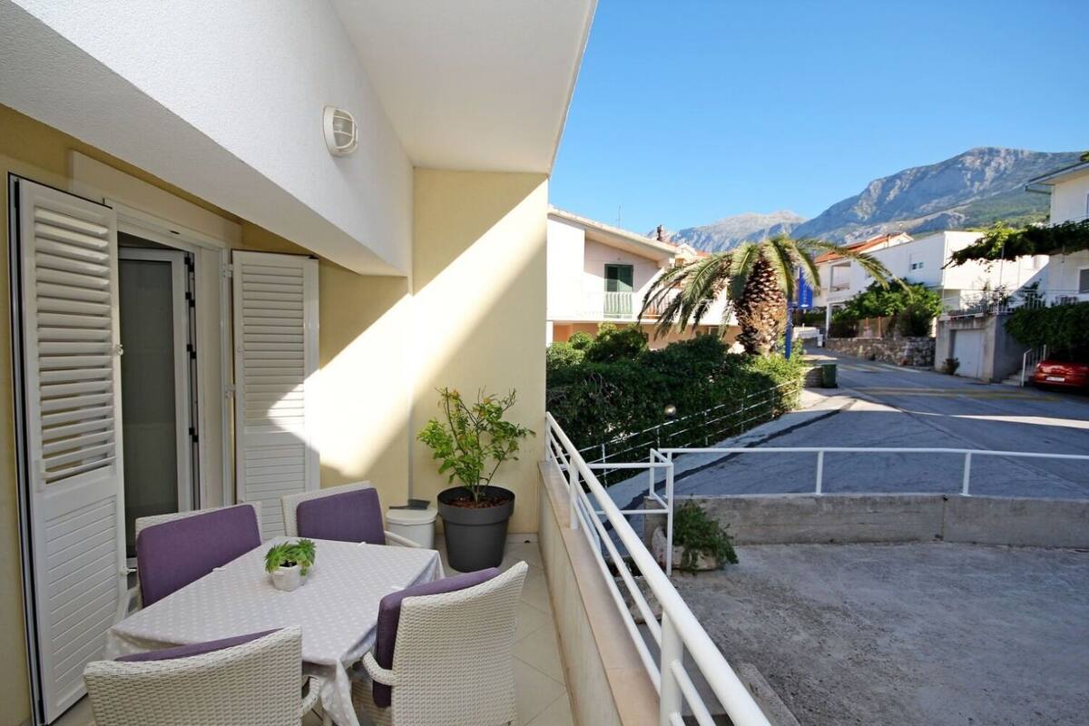 Ein Balkon mit Tisch und Stühlen, einem Blumentopf und Blick auf eine Straße mit Häusern und Bergen im Hintergrund.
