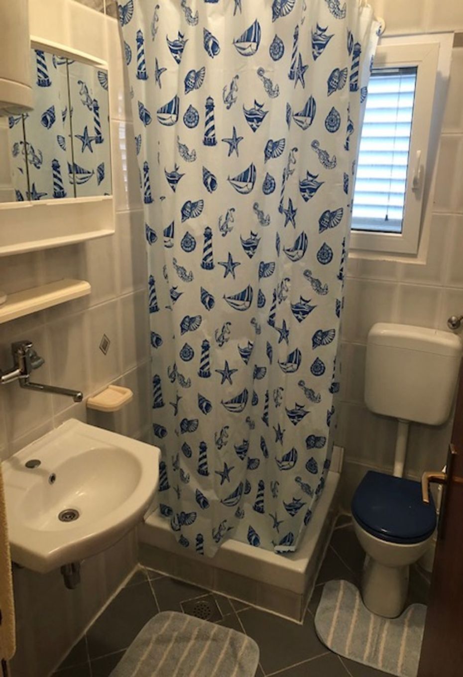 Et badeværelse med bruseforhæng med maritimt tema, toilet, vask og et vindue med persienner.