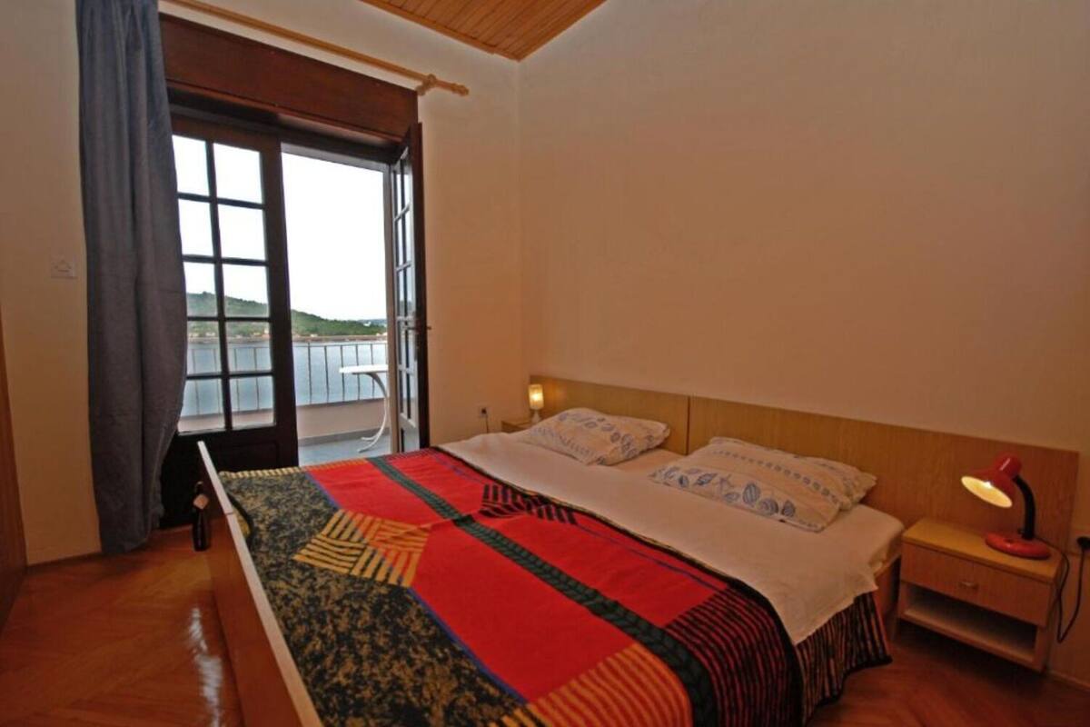 A bedroom with a large bed, a nightstand with a lamp, and a view of a body of water through an open door.