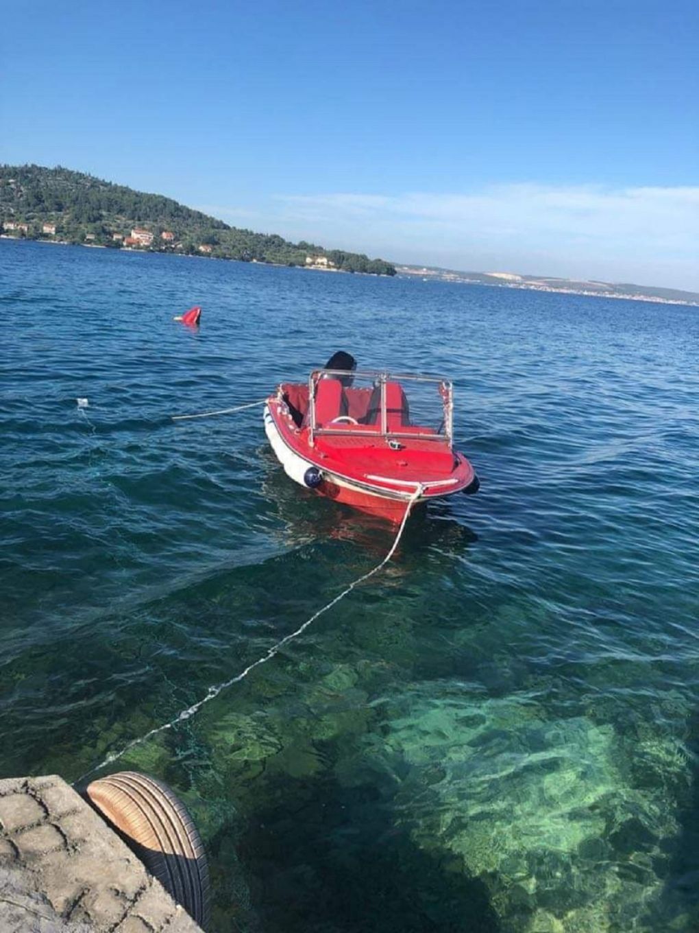 A small red boat tied to a dock with clear water and a distant shoreline.