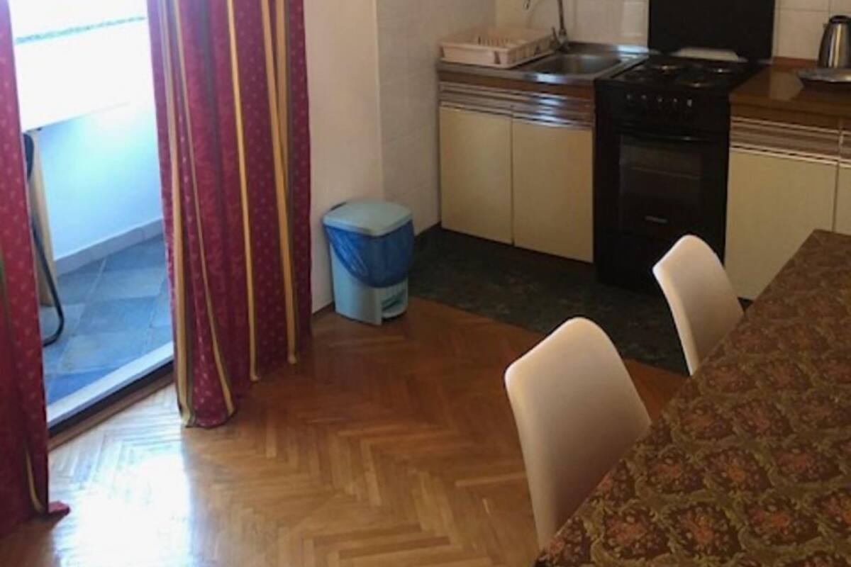 A compact kitchen with a sink, stove, and cabinets. There is a dining table with chairs and a trash bin.