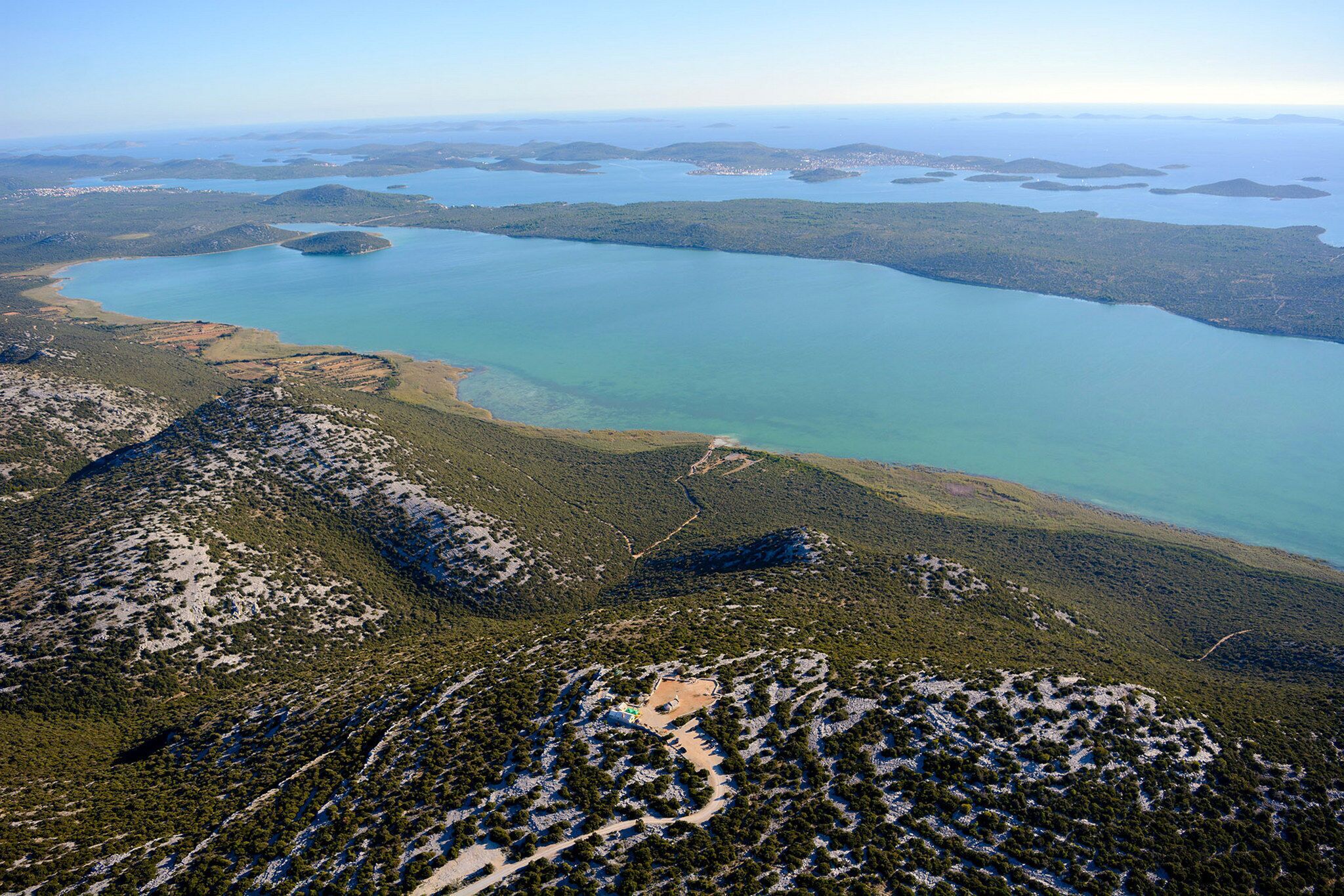 An aerial view of a coastal landscape with a large body of water, surrounded by forested hills and a winding road.