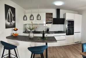 A modern kitchen with white cabinets, a central island with bar stools, pendant lights, and a black and white artwork on the wall. - House Noa (Orebic)