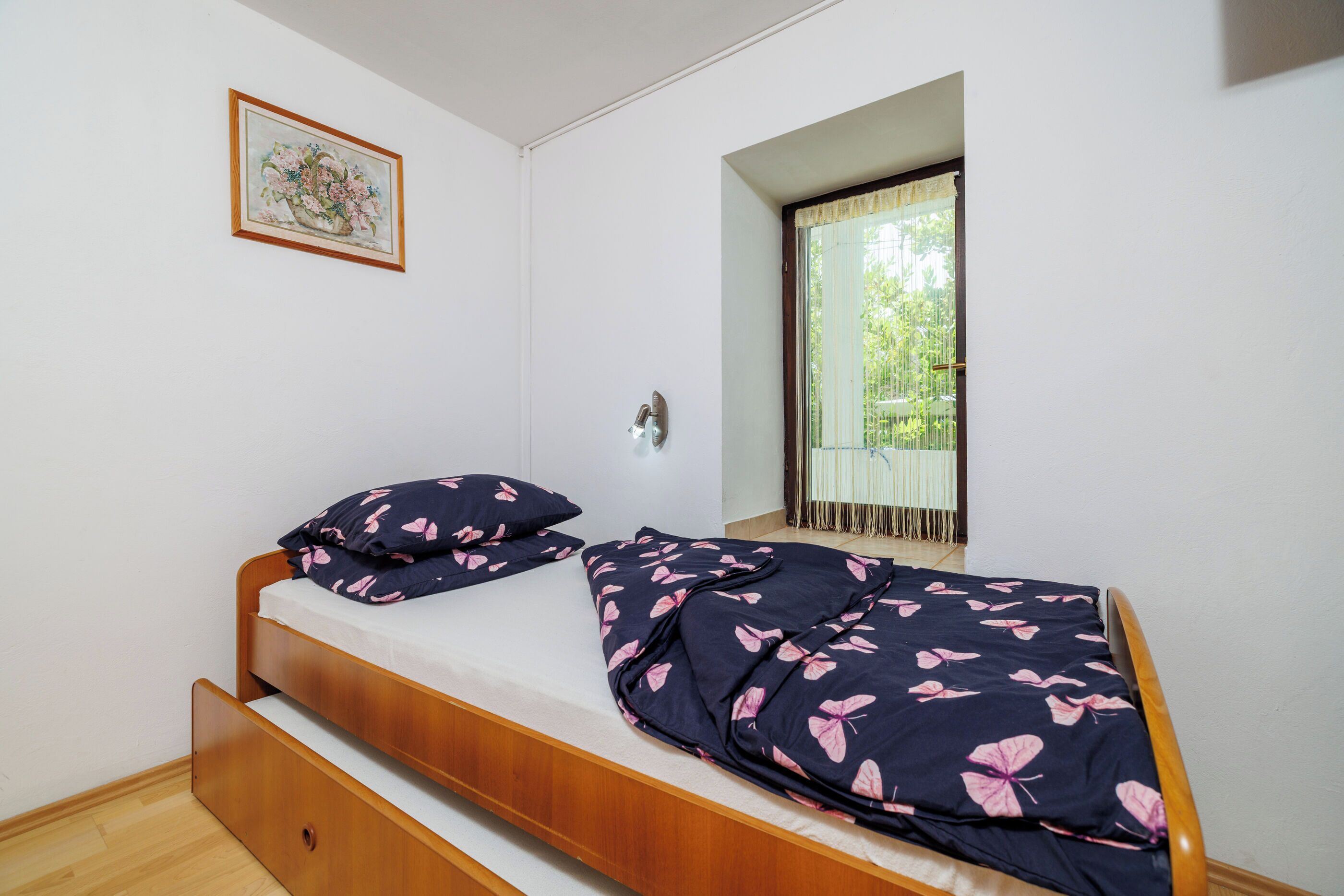 Un letto singolo con struttura in legno, una finestra vista su spazi verdi e un quadro appeso al muro.