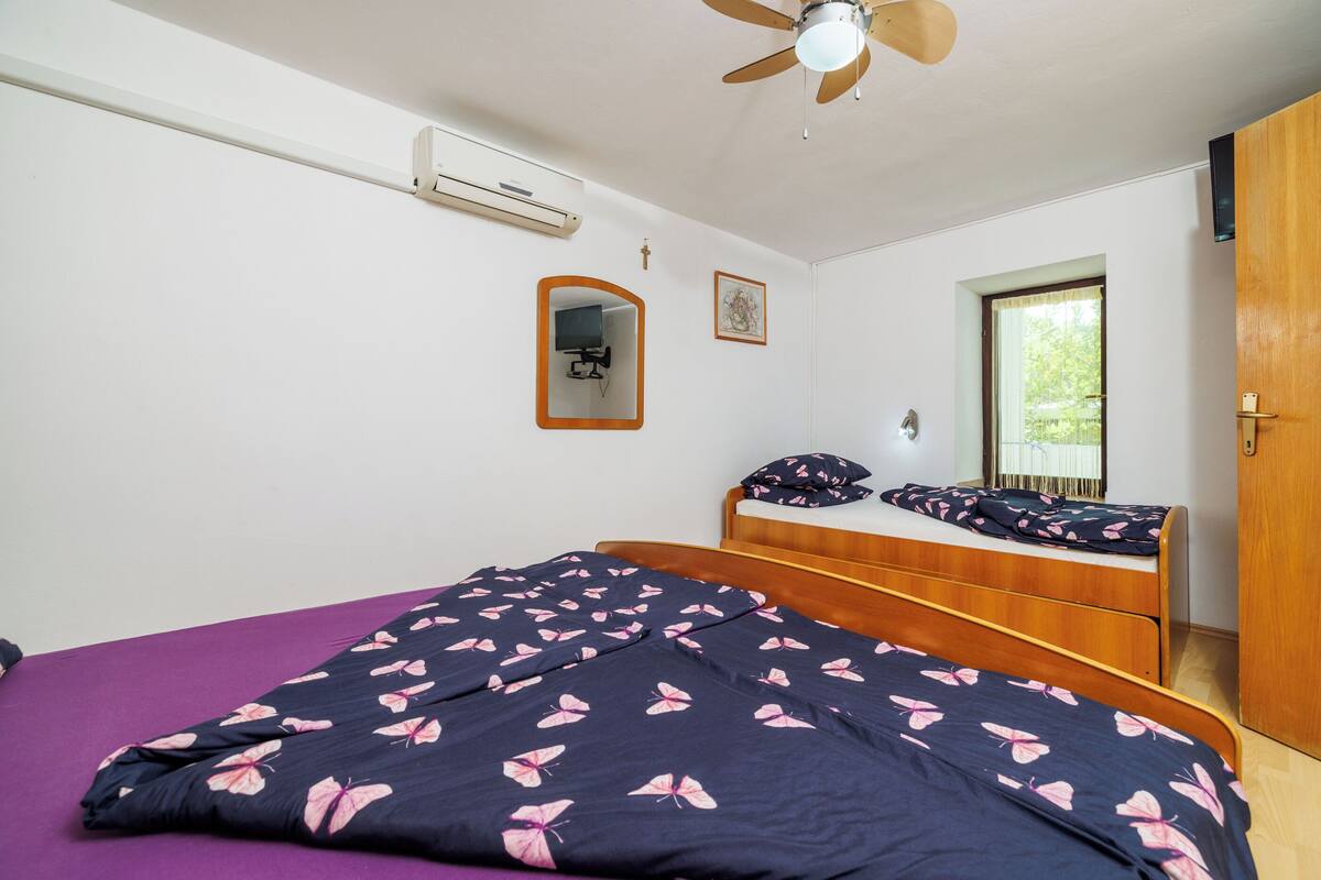 Une chambre à coucher comprenant un lit, un ventilateur de plafond, un appareil de climatisation, une armoire en bois, une fenêtre avec vue et un tableau encadré accroché au mur.