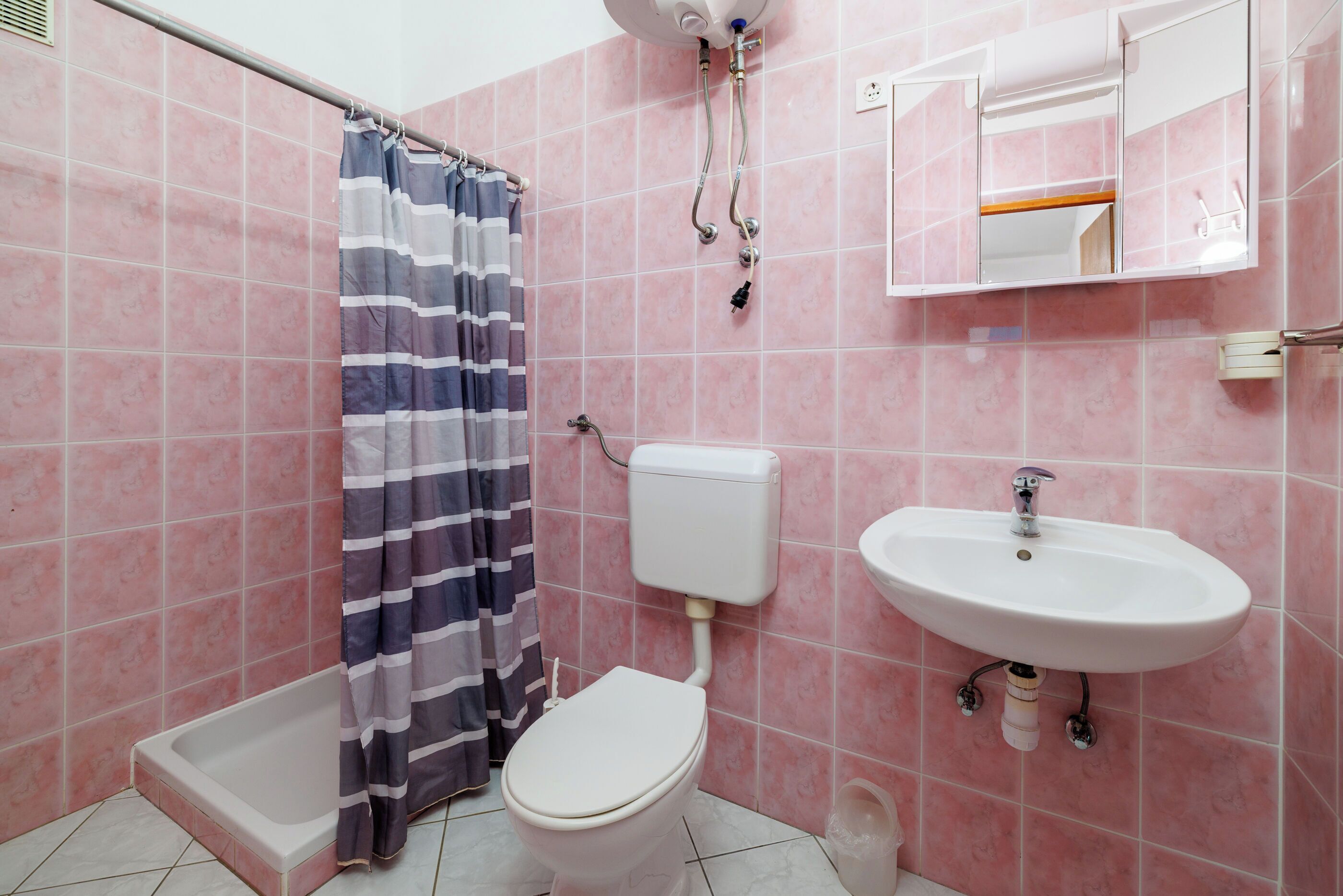 Een badkamer met roze betegelde muren, een wit toilet, een douche met een gestreept gordijn, en een wit wastafeltje met een spiegel erboven.