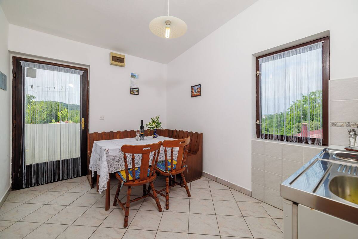 Un coin repas avec une table et des chaises, une fenêtre avec des rideaux, un luminaire au plafond et un espace cuisine avec un évier.