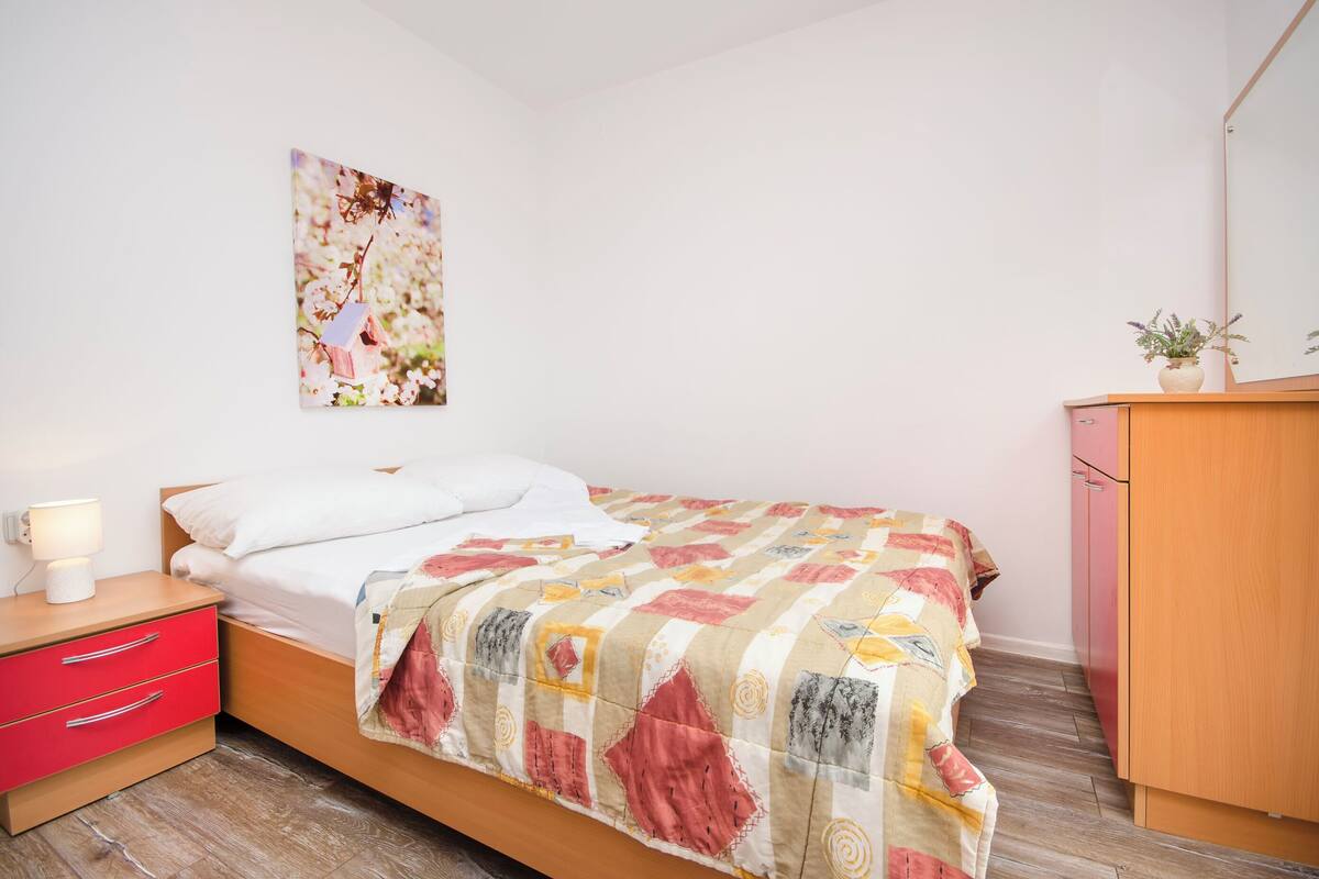 Een netjes opgemaakt bed met een kleurrijke sprei, een nachtkastje met een lamp, een rode lade en een houten garderobe.
