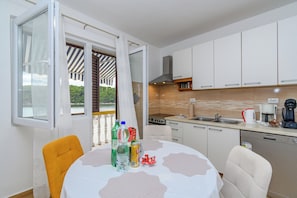 A modern kitchen with white cabinets, a stove, and a sink. There is a dining table with chairs, and a window with a view of greenery. - Apartments Ivi (Rab)