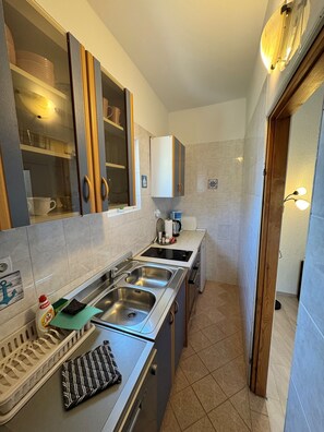 A compact kitchen with a double sink, a stove, and a small countertop area. There are cabinets above the sink and a window with a view of the outside. - Apartments Ivi (Rab)