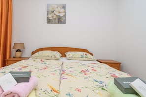 A double bed with floral bedding, a bedside lamp, a painting on the wall, and a 'Welcome' box on the nightstand. - Apartments Ivi (Rab)