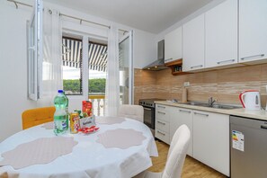 A modern kitchen with white cabinets, a stove, and a sink. There's a table with a white tablecloth and a bottle of water. - Apartments Ivi (Rab)