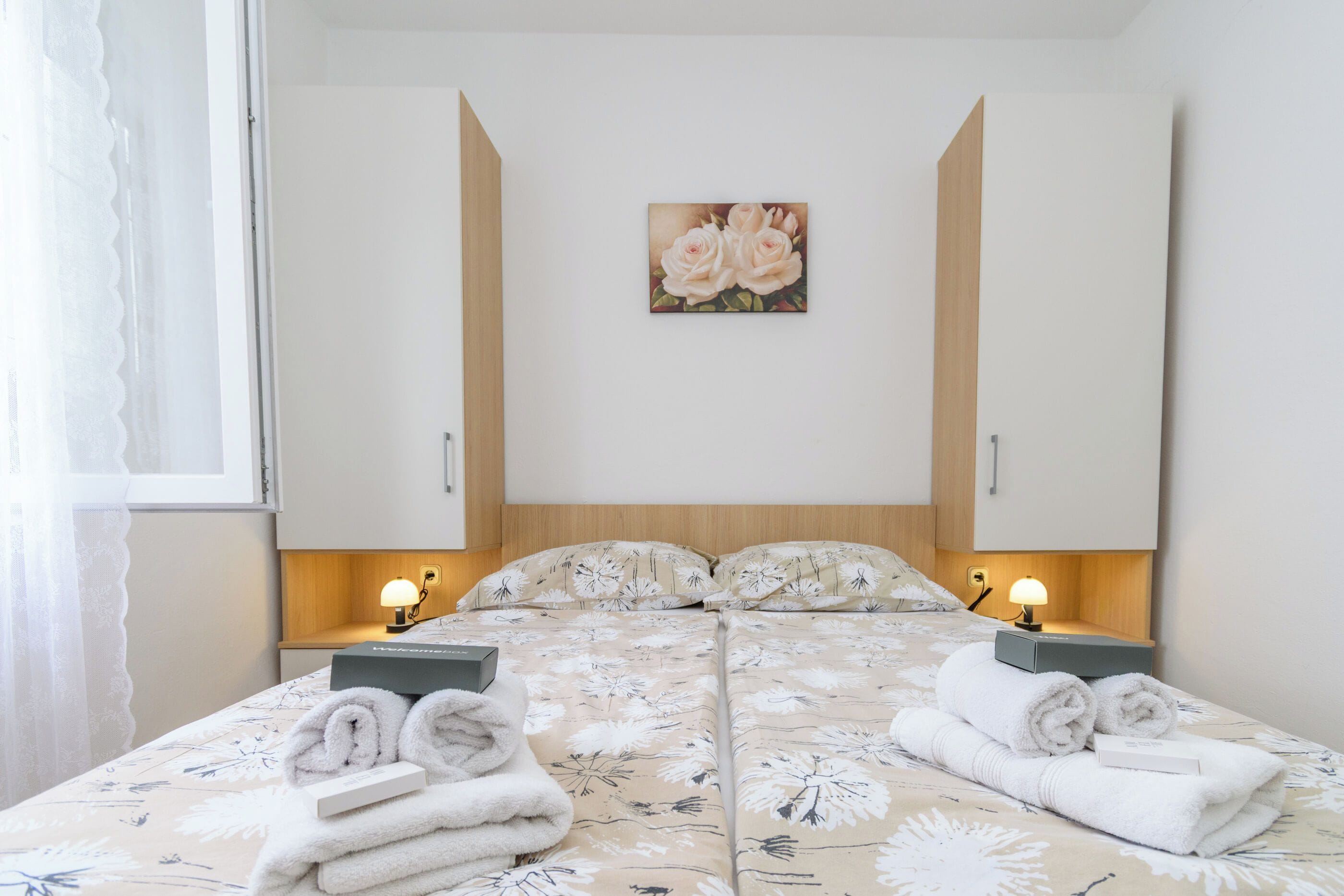 Un lit double avec une parure de lit à motifs floraux, deux lampes de chevet, un tableau représentant des roses et une boîte contenant des articles de toilette.
