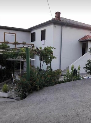A white building with a red-tiled roof, a green pergola, and a small garden area with plants and a white chair. - Apartments Ivi (Rab)