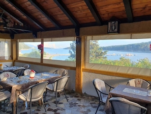 Un área de estar al aire libre techada con mesas y sillas, ventanas amplias con vista a un lago y barcos, y techo de madera.
