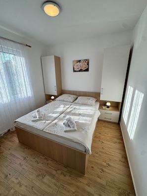 A bedroom with a wooden bed, white bedding, a nightstand with a lamp, a wardrobe, and a painting on the wall. - Apartments Ivi (Rab)