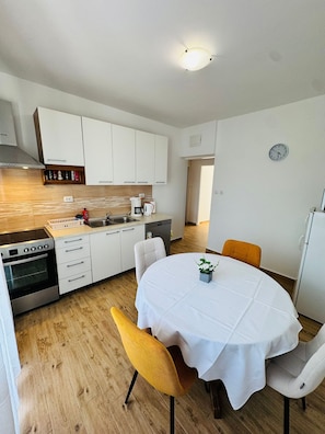 Una cocina compacta con armarios blancos, horno empotrado y un lavamanos. Hay una mesa de comedor con mantel blanco y dos sillas.