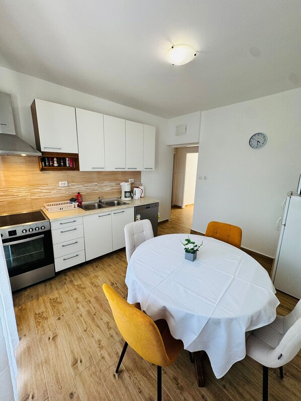 A compact kitchen with white cabinets, a built-in oven, and a sink. There is a dining table with a white tablecloth and two chairs. - Apartments Ivi (Rab)