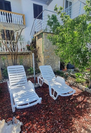 Two white lounge chairs in a garden area with a stone wall and a tree. - Apartments Ivi (Rab)