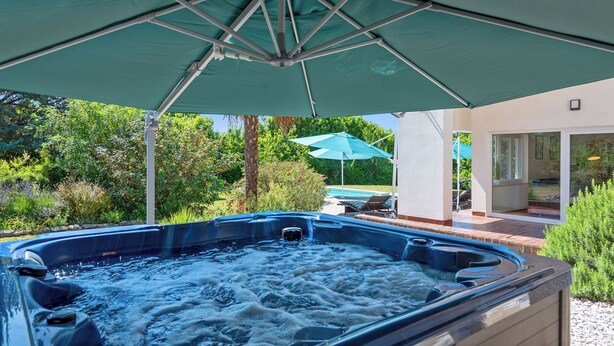 Una vasca idromassaggio sotto un ombrellone verde, immersa nel verde e in prossimità di una piscina.