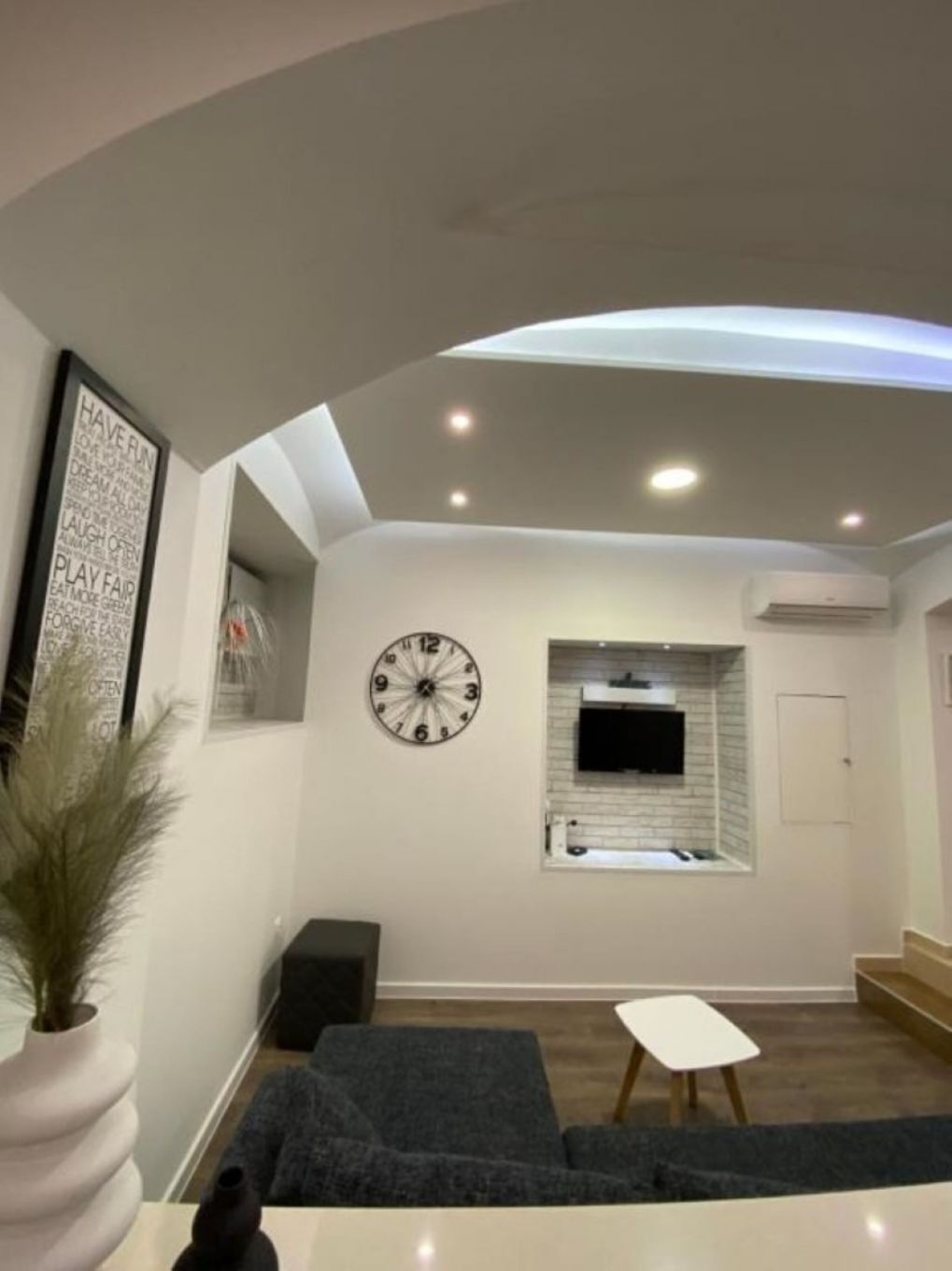 Un salon moderne avec un canapé gris, une table basse blanche, une horloge murale et un téléviseur à écran plat fixé au-dessus d’une cheminée.