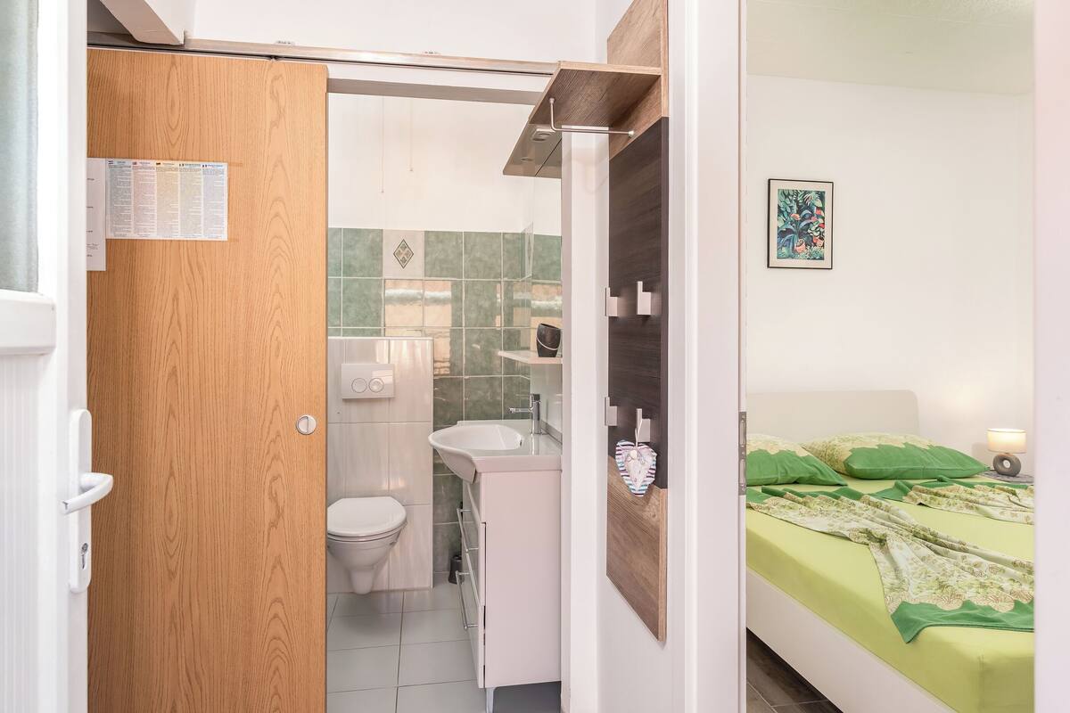 A room with a wooden door, a bathroom with a toilet and sink, and a bed with green bedding.