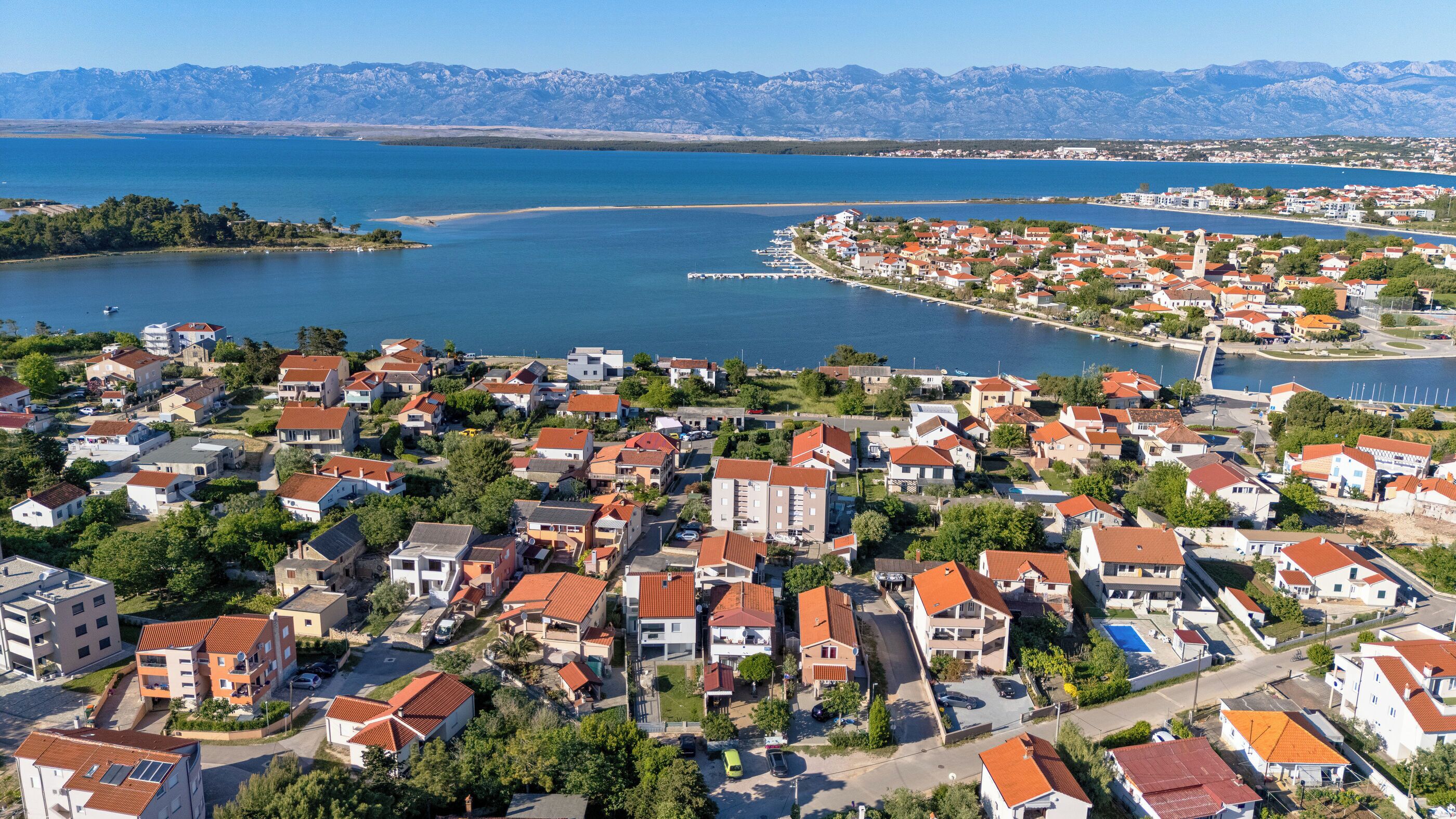 A coastal town with residential buildings, a marina, and a mountainous backdrop.