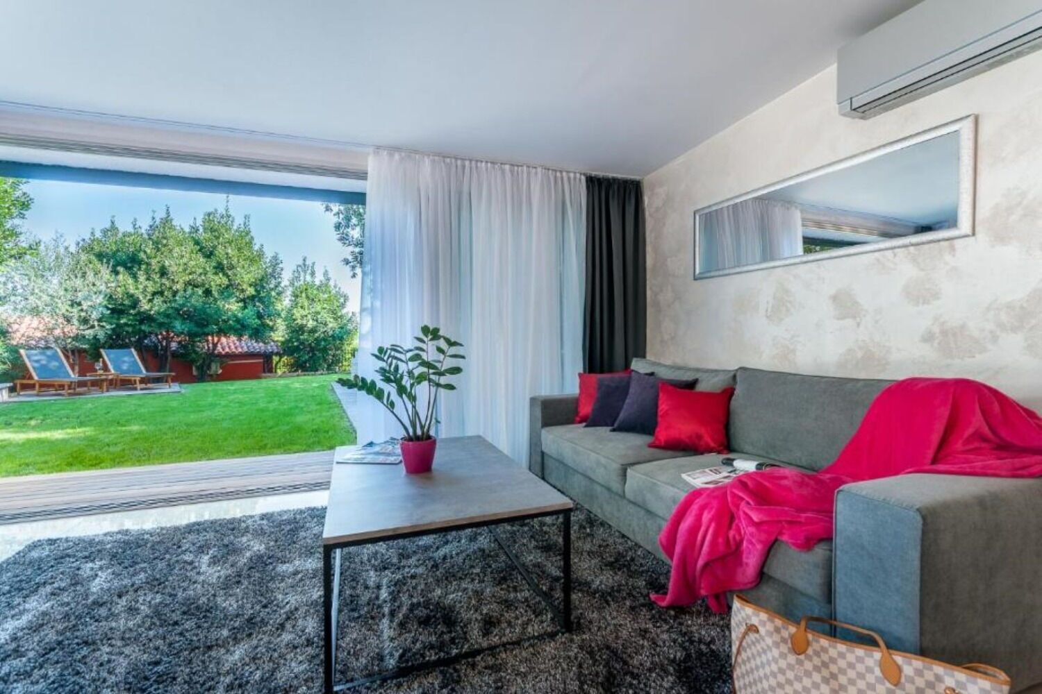 A modern living room with a grey sofa, a coffee table, and a potted plant. There is a large window with curtains, a view of a garden with outdoor furniture, and a wall-mounted air conditioning unit.