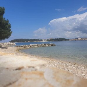A coastal scene with a rocky shoreline, clear water, and a distant island with buildings. - Apartments Ivo (Rovinj)