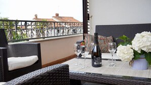 Un balcón con muebles de mimbre, una mesa con copas de vino y una botella, y flores blancas.