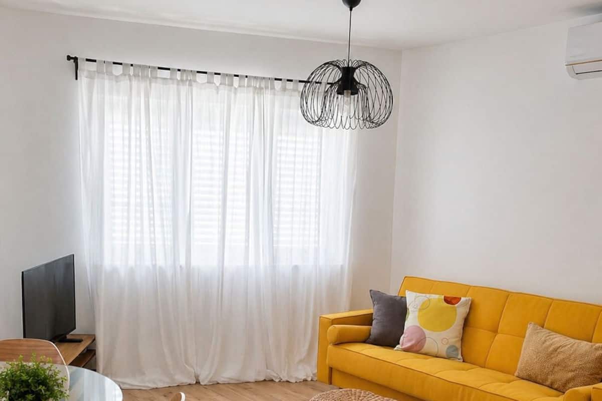 A modern living room with a yellow sofa, a glass dining table, and a wicker ottoman.